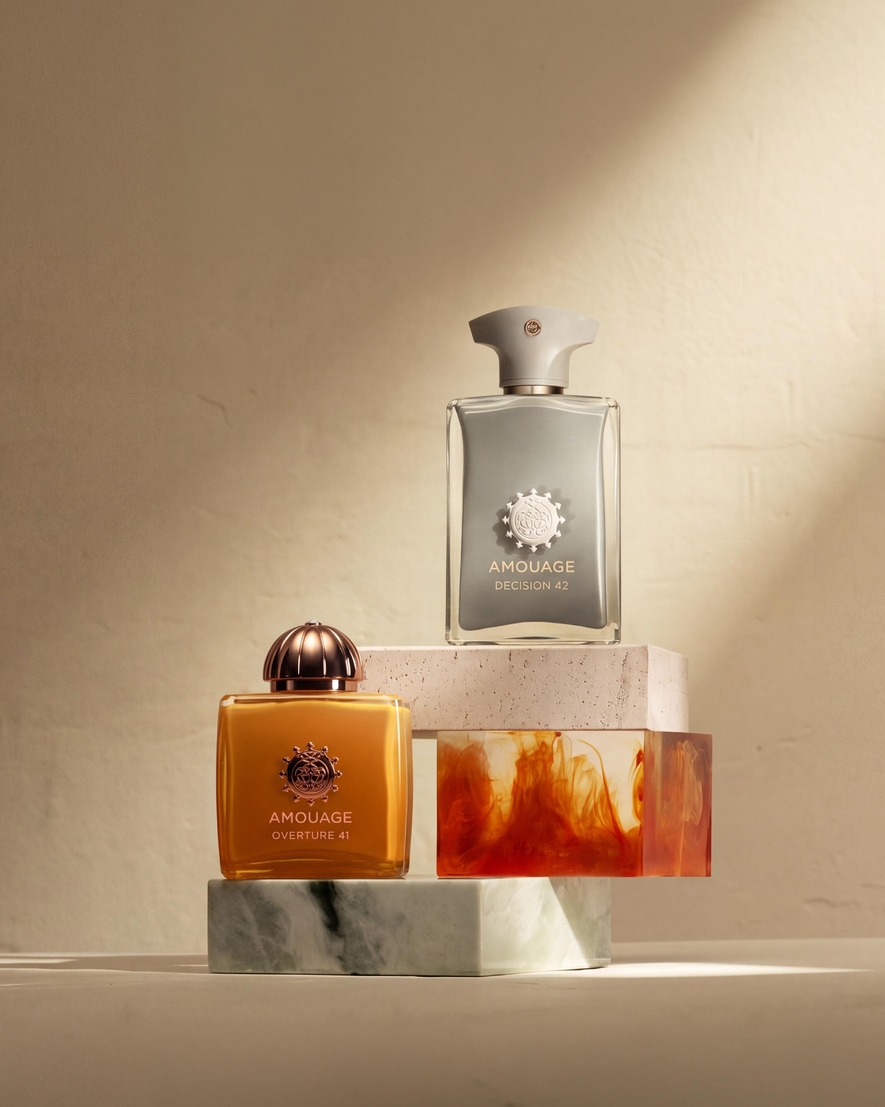 A grey bottle of Amouage Decision 42 above an amber Overture 41 bottle on sculptural blocks in warm light.