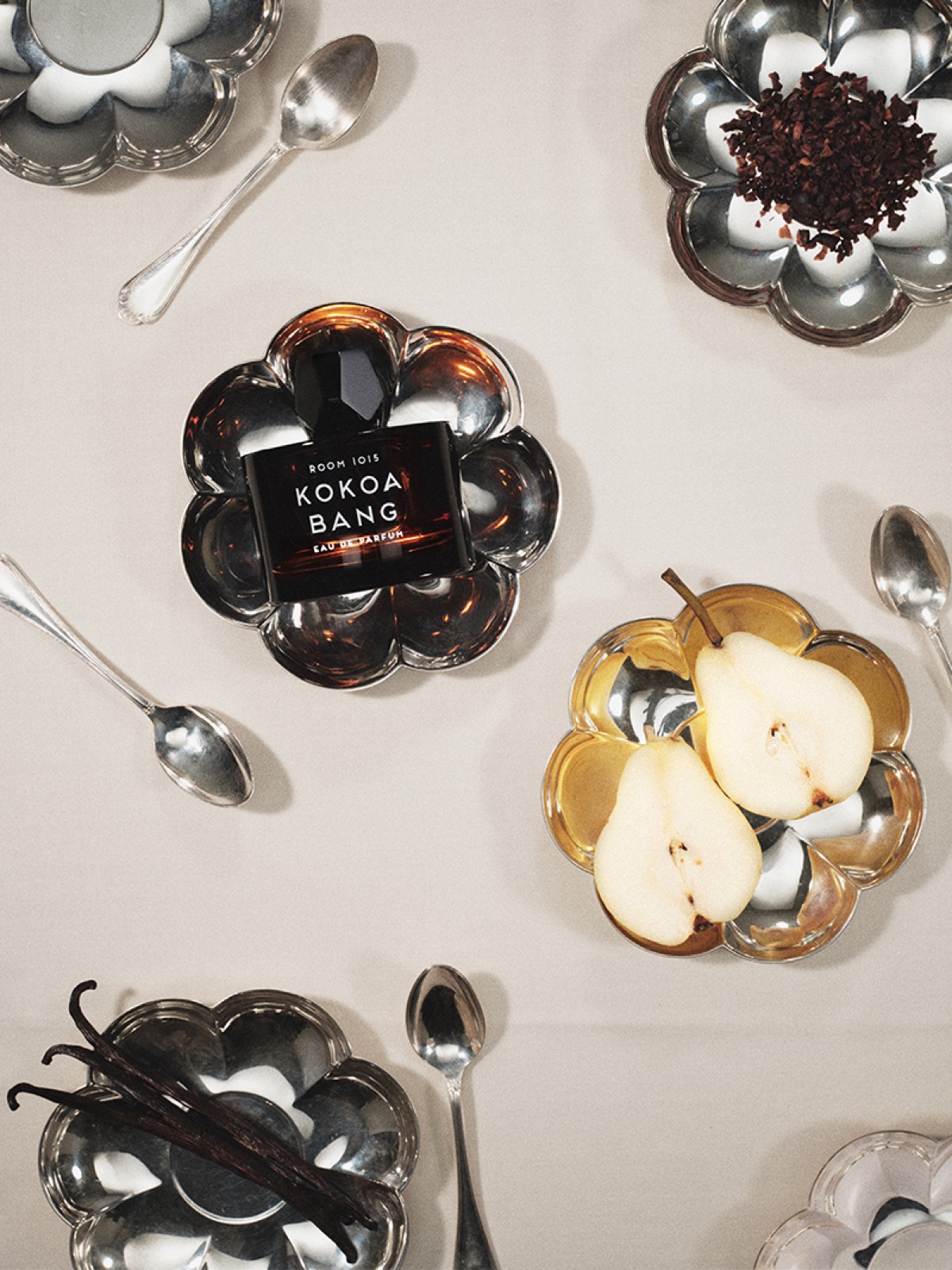 A dark amber Room 1015 Kokoa Bang bottle surrounded by pears, cocoa nibs, vanilla pods and silver spoons.